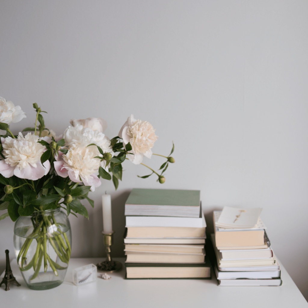 Stack of pretty books to the right with a green vase of soft white flowers to the left.